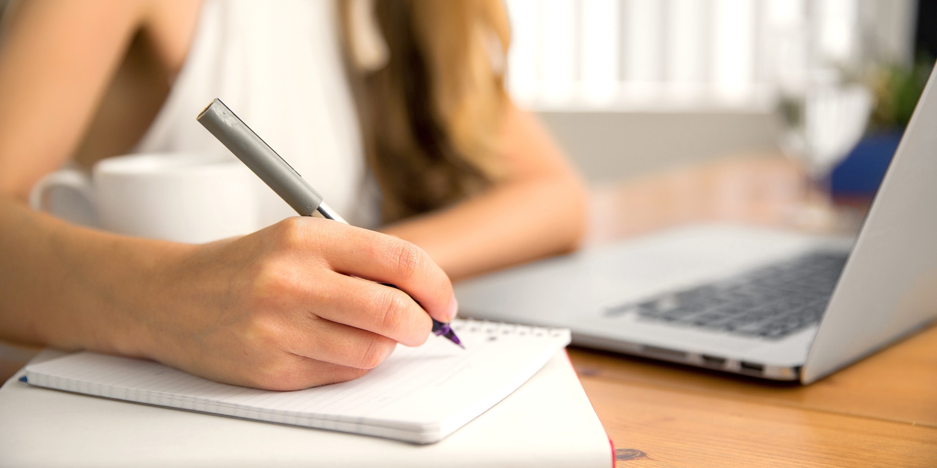 Woman writing in a notepad next to a laptop on a desk