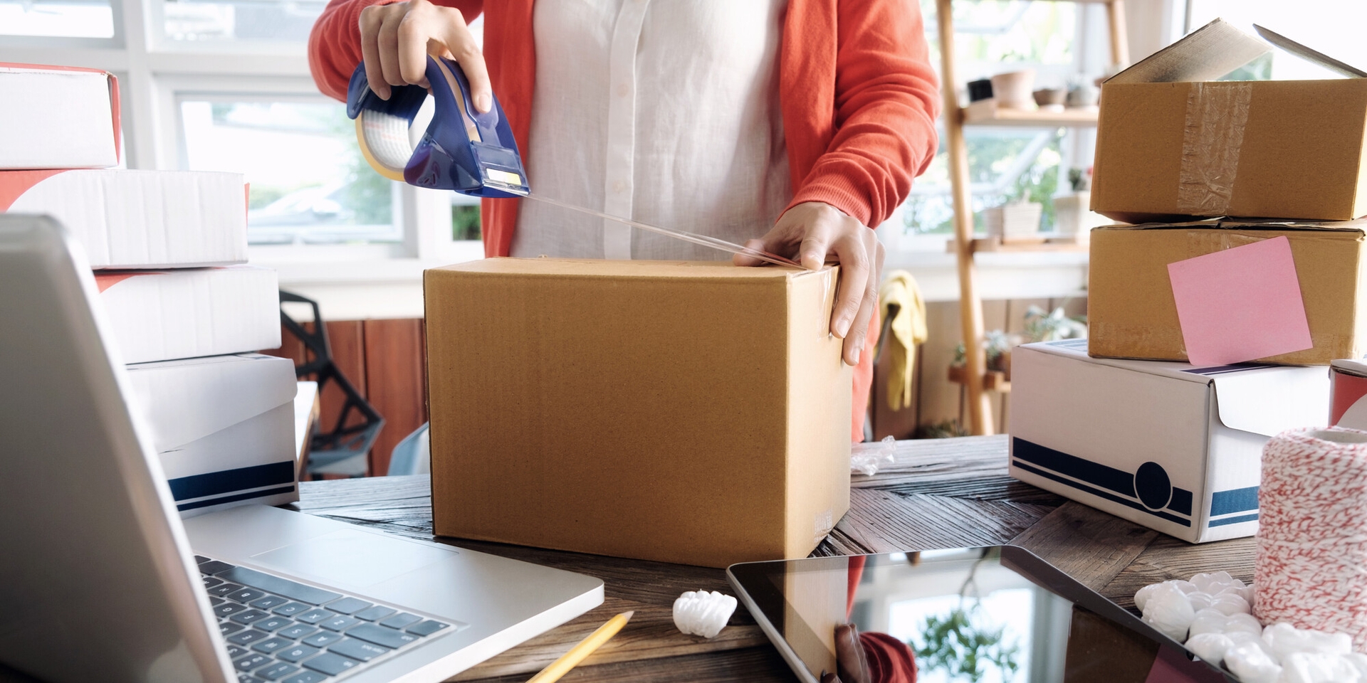 Person taping a cardbox box closed on a desk infront of a laptop and other boxes.